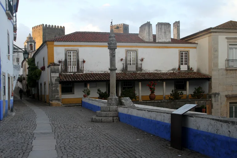 The stone pillory on its stepped base at the top of the Praça de Santa Maria, before a long white house with an arcaded ground floor and castle towers behind.