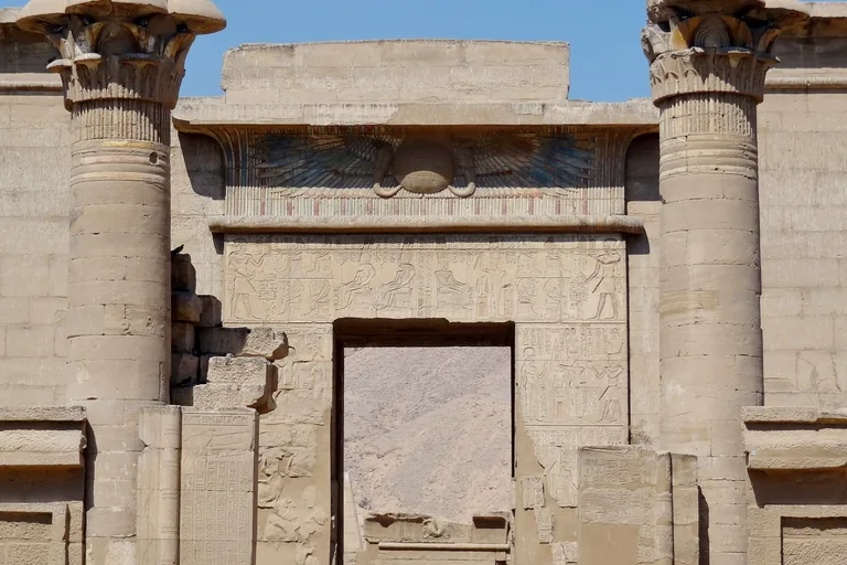 The late stone gateway of the small temple of Amun at Medinet Habu, with a painted winged disk over the door between two tall columns with carved capitals and the desert hills seen through the doorway.