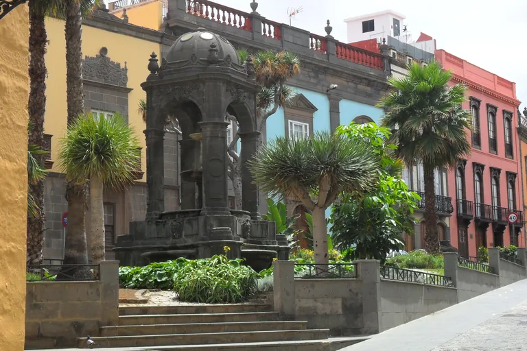 A domed stone pavilion with arches over the fountain in the Plaza del Espíritu Santo, in a raised garden of dragon trees and palms reached by steps, with house fronts in yellow, blue and red behind.