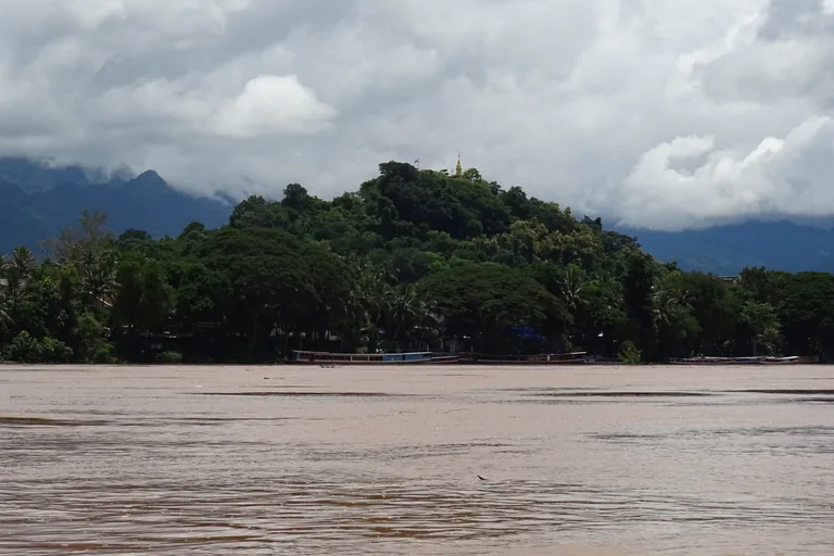 The wooded hill of Phousi in Luang Prabang with a small gilded spire on its summit, seen across the wide brown Mekong, with long boats moored along the bank and mountains in low cloud behind.