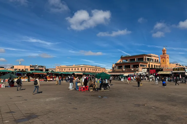 The wide paved Jemaa el Fna under a blue sky, with people walking among red stools, juice stalls under green awnings and a minaret rising over the buildings on the right.