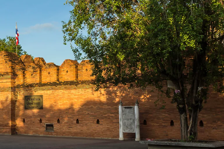 The red brick wall of Tha Phae Gate in Chiang Mai with its battlements, a name plaque on the wall, a flag above and a large tree at the right, seen across a wide paved square under a blue sky.