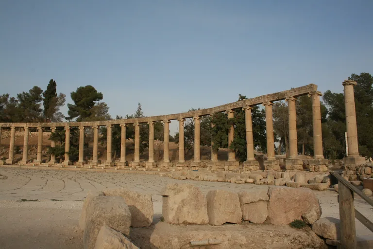 The Oval Plaza at Jerash, a long curving row of Ionic columns carrying their stone beams around the paved square, with trees behind and fallen blocks in the foreground.