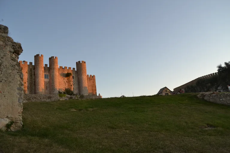 The round and square crenellated towers of the Castle of Óbidos in evening sun beyond a grassy slope inside the walls.