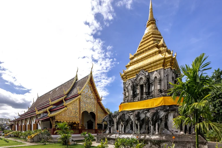 The main hall of Wat Chiang Man in Chiang Mai with its tiered dark roofs and gilded carved gable, and beside it a chedi with a row of grey stucco elephants around its base and a gilded upper part and spire, under a blue sky with white cloud.