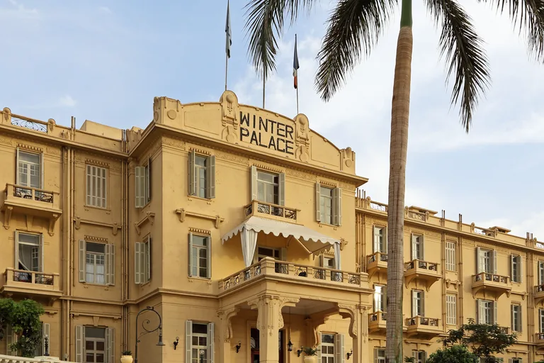 The yellow river front of the Winter Palace hotel in Luxor, with WINTER PALACE lettered on its central pediment, shuttered windows, an awning over a balcony and a tall palm tree in front, under a pale blue sky.
