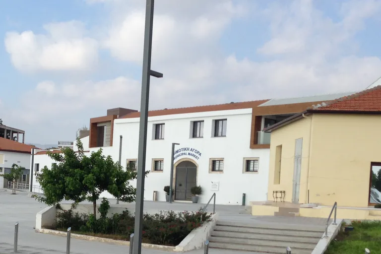 The white market hall of Paphos with an arched doorway under blue lettering naming the Municipal Market in both Greek and English, wood clad rooms on its upper floor, a young tree and steps in front and a pale yellow building at the right.