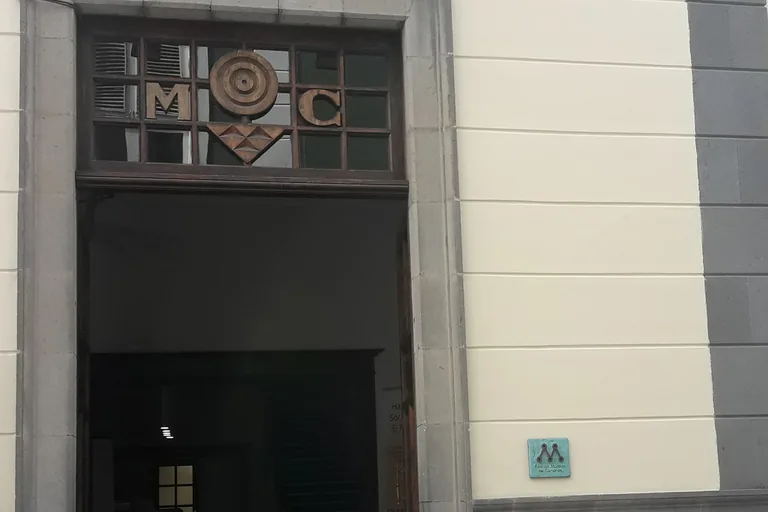 The tall stone framed doorway of El Museo Canario in a cream wall, its wooden fanlight set with the letters M and C round a carved disc, with a small green plaque on the wall beside it.