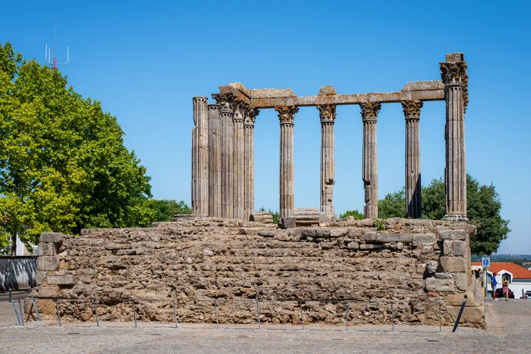 The granite podium and Corinthian columns of the Roman Temple of Évora behind a low fence, under a clear sky.