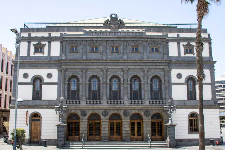 The front of the Teatro Pérez Galdós, grey stone and white walls with five arched doorways at the top of a flight of steps, a row of arched windows above and the theatre's name under the crest, with a street lamp at the left and a tall palm at the right.