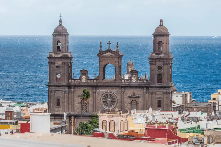 The dark stone front of the Cathedral of Santa Ana with its two domed bell towers and an open arched gable between them, seen over the white and red rooftops of Vegueta with the blue Atlantic behind.