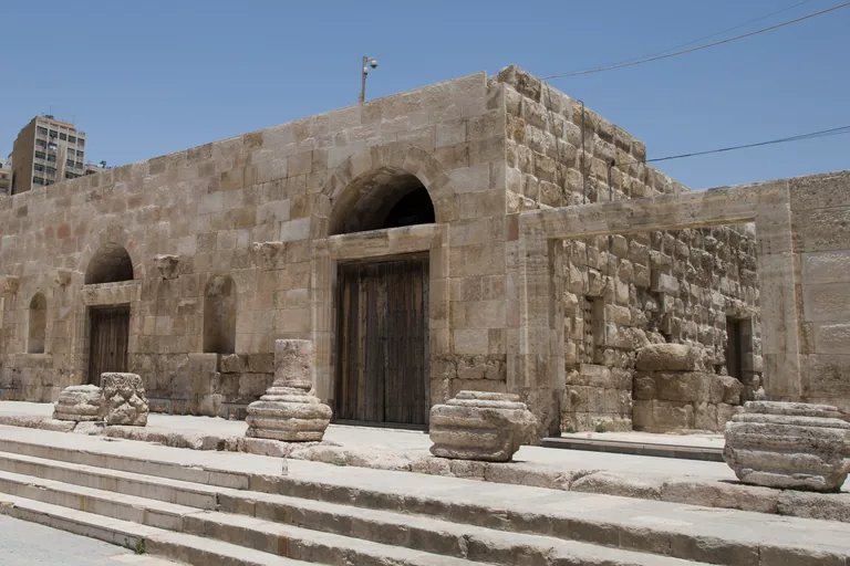 The Odeon in Amman, a restored Roman building of squared stone with tall wooden doors under arches and carved column bases along its terrace above broad steps, with a tall building on the hill behind.