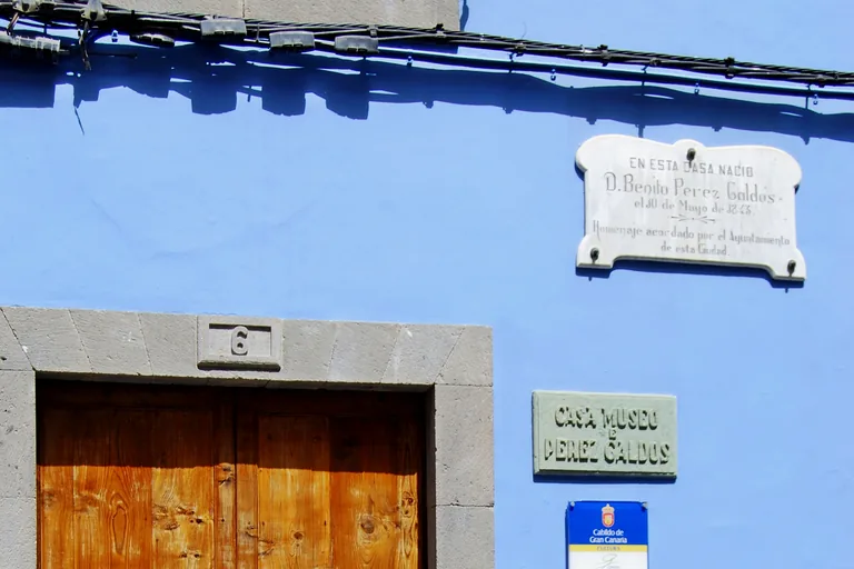 Part of a blue plastered house front in Calle Cano, with a marble plaque recording that Benito Pérez Galdós was born in the house and a green sign reading Casa Museo Pérez Galdós, above the stone frame and the top of a wooden double door.