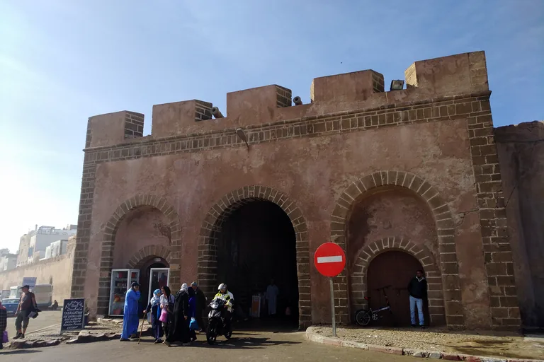 Bab Doukkala, a crenellated gate of rose plaster and cut stone with three arches and small cannons on top, with people and a motorbike passing in front.