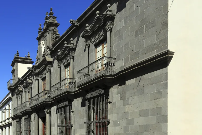 The dark grey stone front of Casa Salazar, with carved window frames, iron balconies on brackets, barred lower windows and a crest high in the middle, seen at an angle along the street.